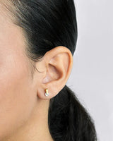 Close-up of an ear wearing a gold earring with a clear gemstone on a neutral background